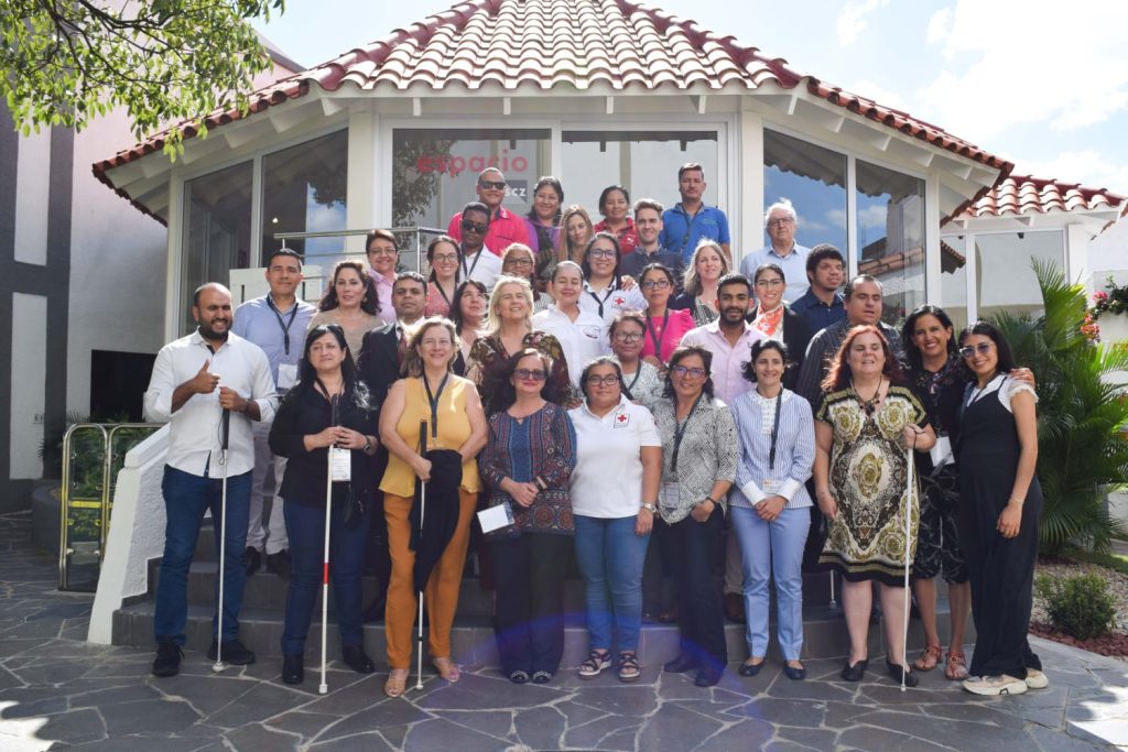 Fotografía de un grupo de aproximadamente 35 o 40 personas con discapacidad visual frente a cámara y detrás de ellos una casa en forma de cabaña con tejas naranja.